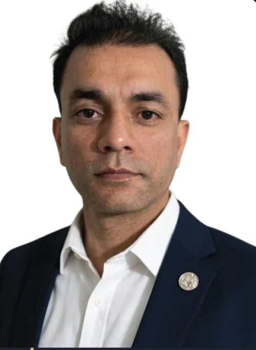 Portrait headshot of a man with dark hair wearing a navy blazer, white shirt, and a circular pin on the lapel against a white background.